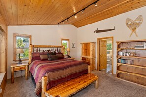A bedroom with a wooden bed, a bench, a bookshelf, and a window with a view of trees.