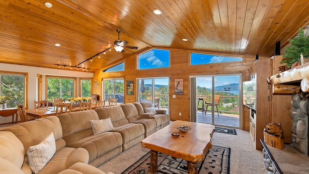 A spacious living room with a large sectional sofa, a wooden coffee table, and a fireplace. There are multiple windows offering views of the outdoors.