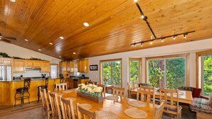 A spacious kitchen with wooden cabinets, a dining area with a large wooden table and chairs, and large windows offering views of greenery.
