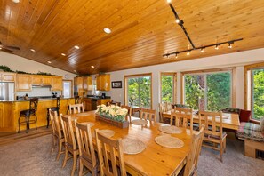 Une cuisine spacieuse avec des meubles en bois, un coin repas doté d’une grande table et de chaises en bois, et de grandes fenêtres offrant une vue sur la verdure.