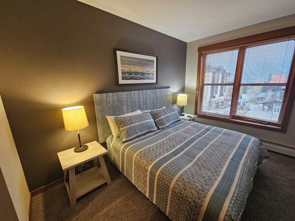 A bedroom with a bed, two bedside tables with lamps, a framed picture on the wall, and a window with blinds. - Copper One 4th Floor—New Carpet, New Furniture, 2 King Beds (Copper Mountain)