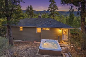 A cabin with a hot tub in front, surrounded by trees and a mountain view.