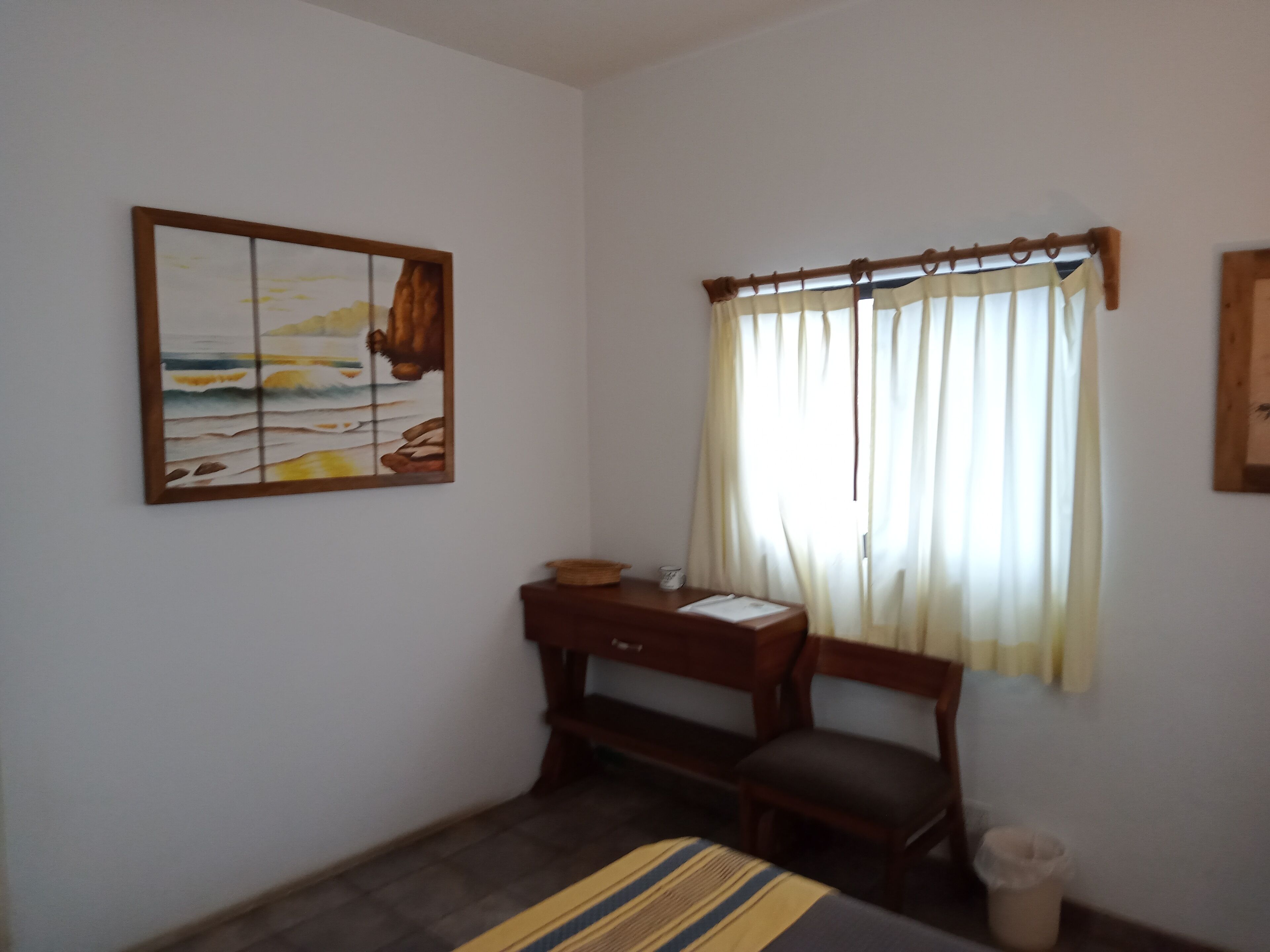 A room with a bed, a desk with a chair, a small table with a basket, a waste bin, and a painting on the wall.