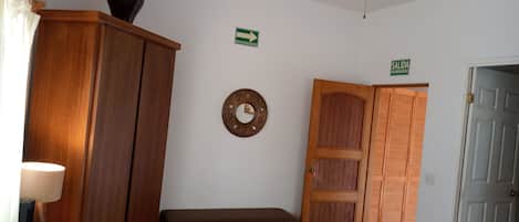 Een hotelkamer met een bed, een houten garderobe, een bankje, een wandklok en een bord met daarop een vluchtroute.