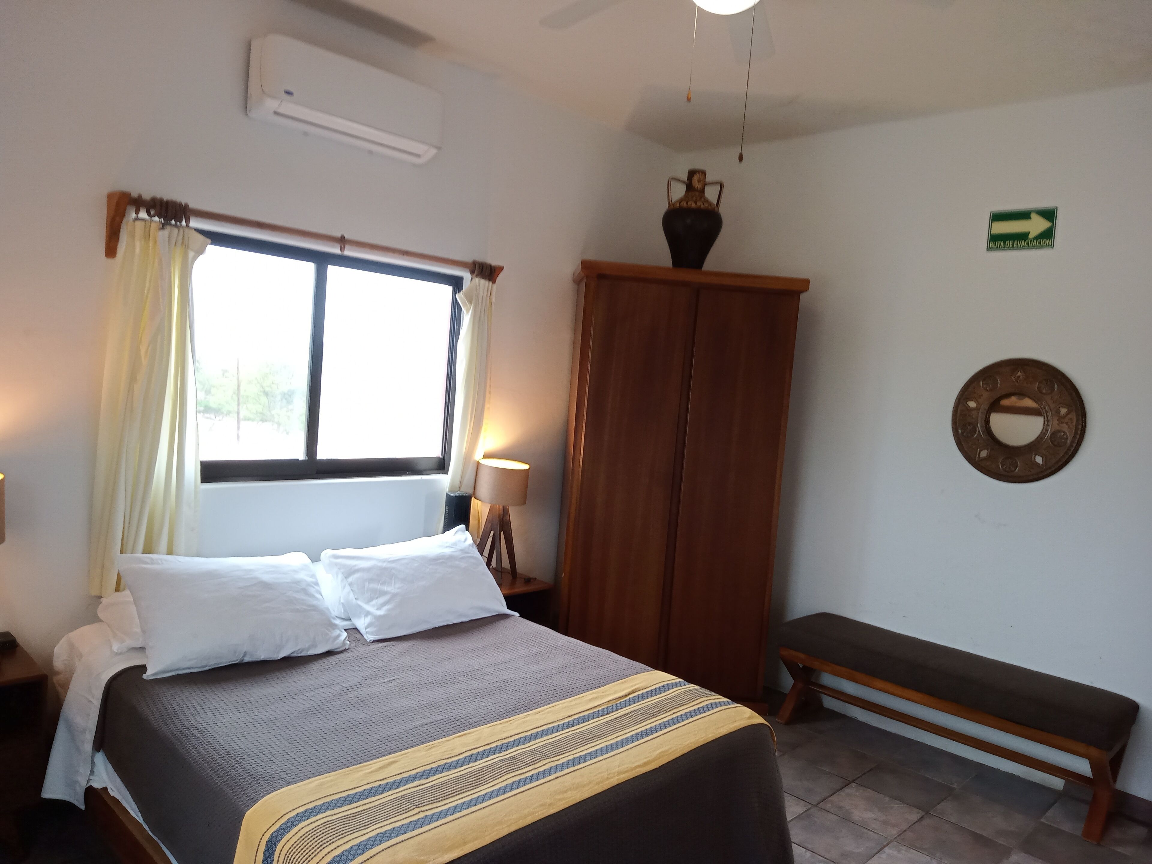 A bedroom with a bed, a bench, a wardrobe, a lamp, and a window with curtains.