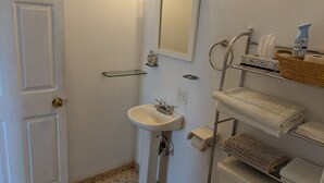 Un baño con lavamanos blanco, espejo e inodoro. Hay un perchero con toallas y una cesta.