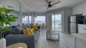 A modern living room with a grey sofa, a dining table, and a kitchen area with a refrigerator.