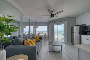 A modern living room with a grey sofa, a dining table, and a kitchen area with a refrigerator.