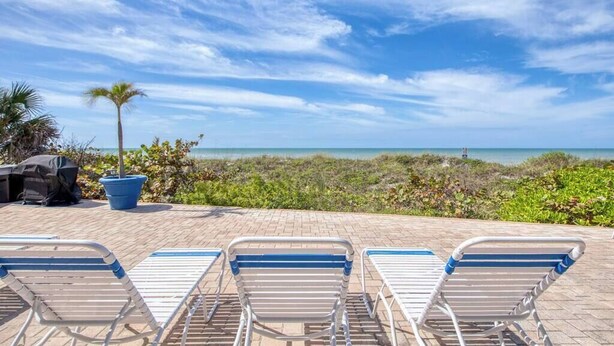 A beachside view with lounge chairs, a grill, and a palm tree.