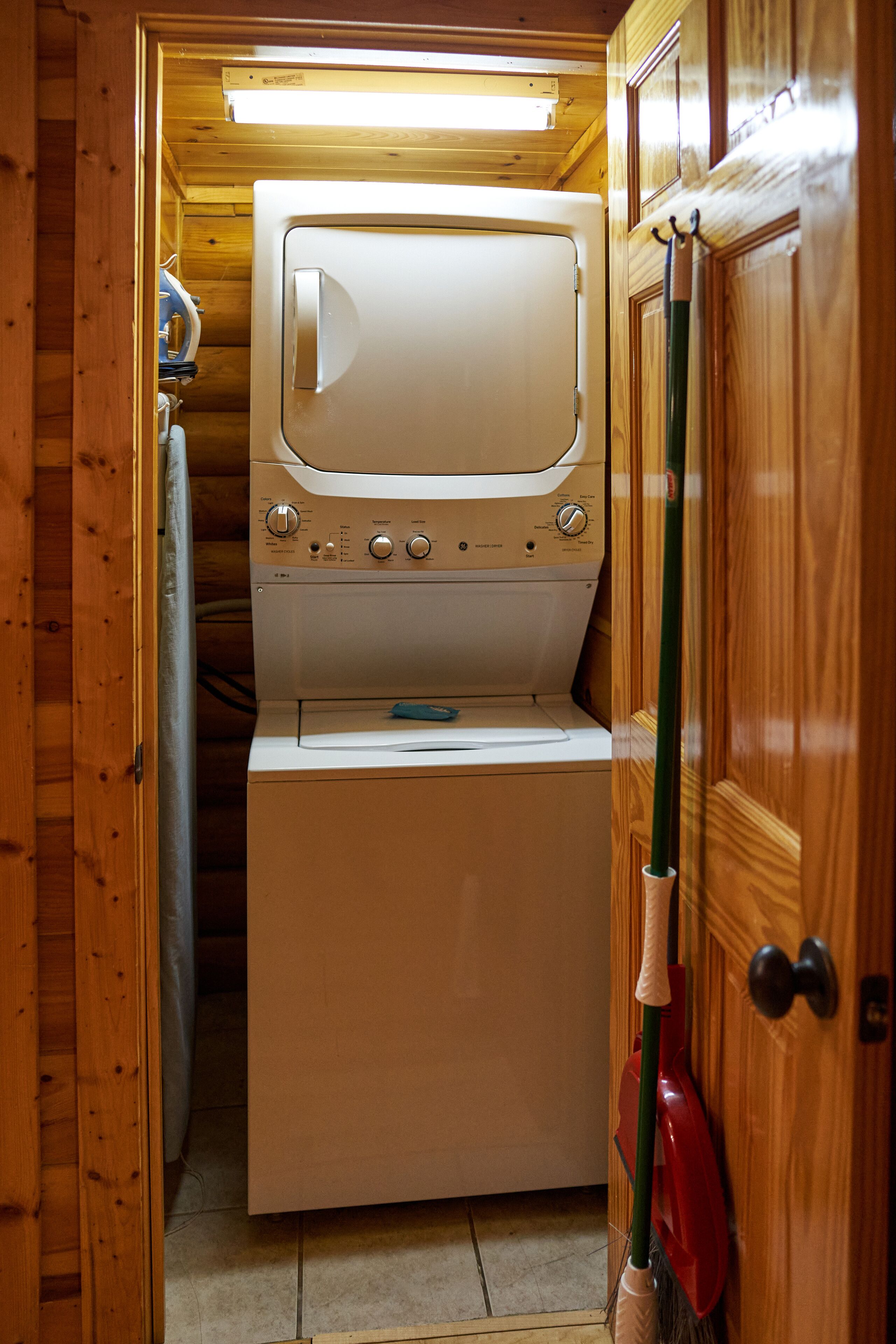 A compact laundry room with a washing machine and dryer, wooden walls, and a broom leaning against the wall.