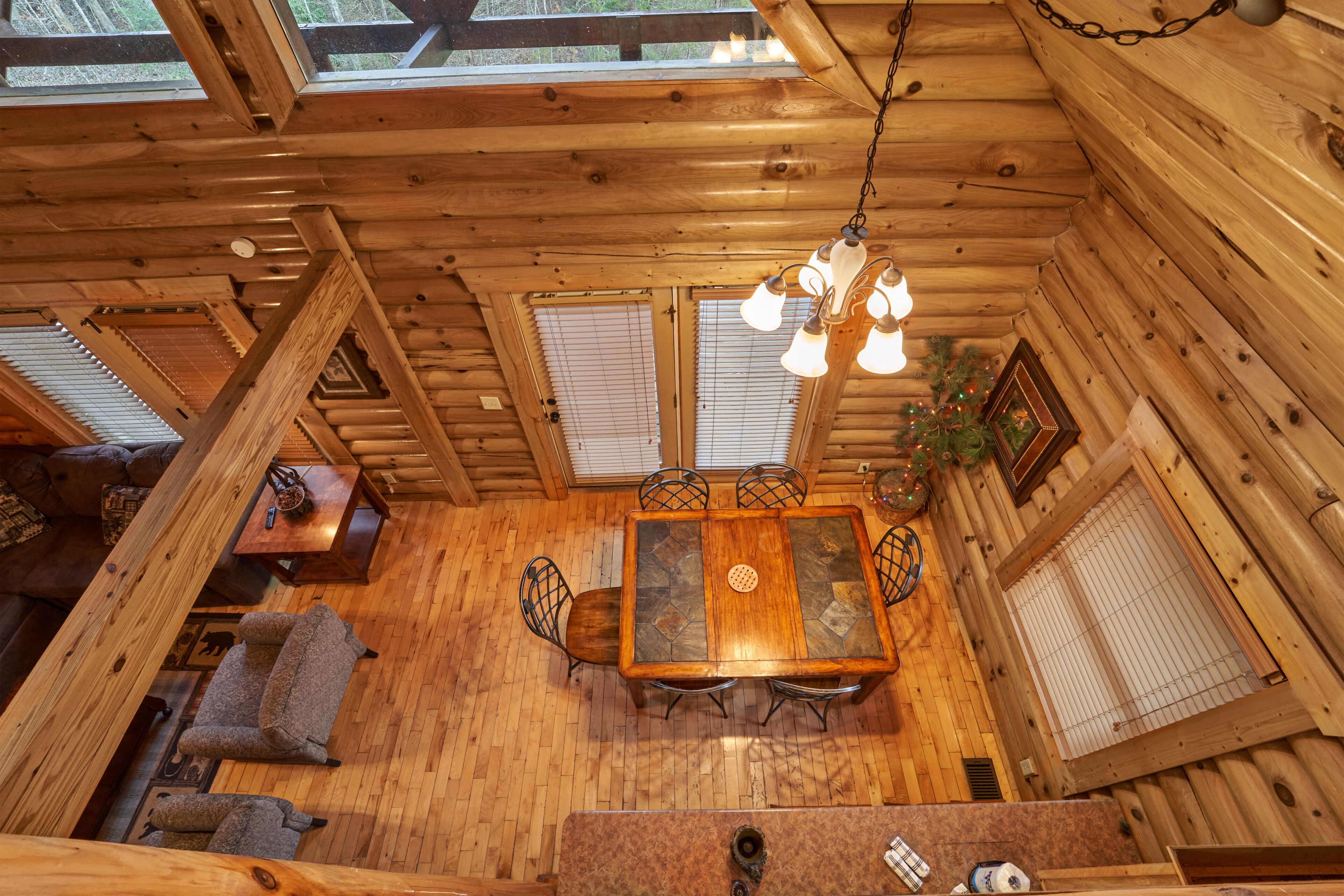 Interior de una cabaña de madera con una mesa de comedor, sillas, una lámpara colgante y un pequeño árbol de Navidad.