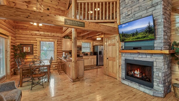 A log cabin interior with a stone fireplace, wooden dining table, and a television mounted above it.