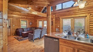 A log cabin interior with a wooden floor, a sofa, a dining table, and a kitchen area with a sink and dishwasher.