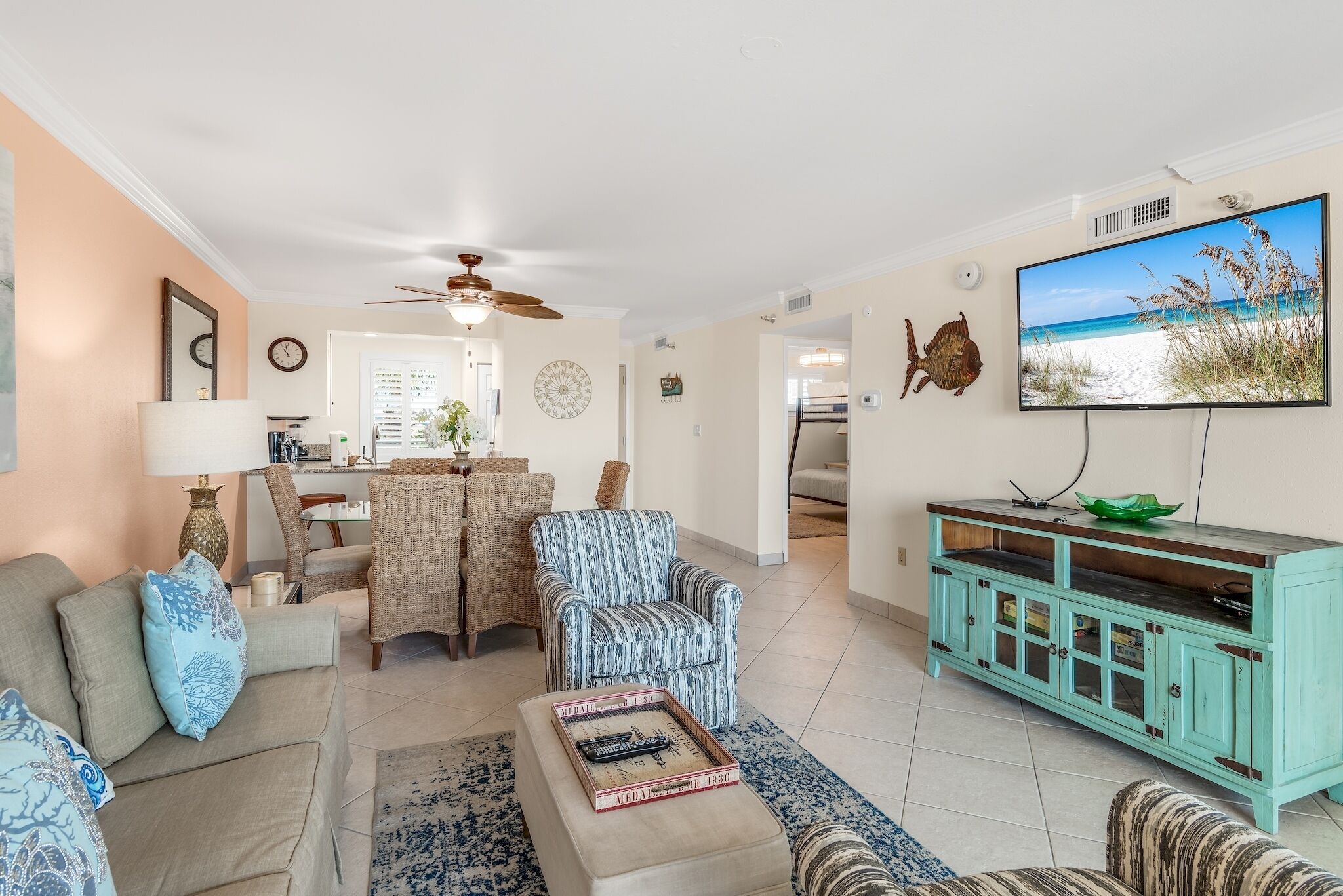 A living room with a sofa, armchair, and ottoman, a dining area with wicker furniture, a teal cabinet, and a TV displaying a beach scene.
