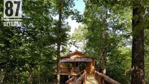 Une cabane en bois dotée d’un porche abrité, entourée d’une forêt dense. Un pont en bois mène à la cabane.