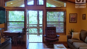 Une pièce chaleureuse avec un plafond en bois, une table à manger avec des chaises, un fauteuil en cuir, un canapé et une grande fenêtre avec des stores.