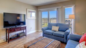A living room with a television, a blue sofa, a wicker ottoman, and a view of the outdoors. - Poolside, spacious and bright! Community boardwalk access to beach (Port Aransas)