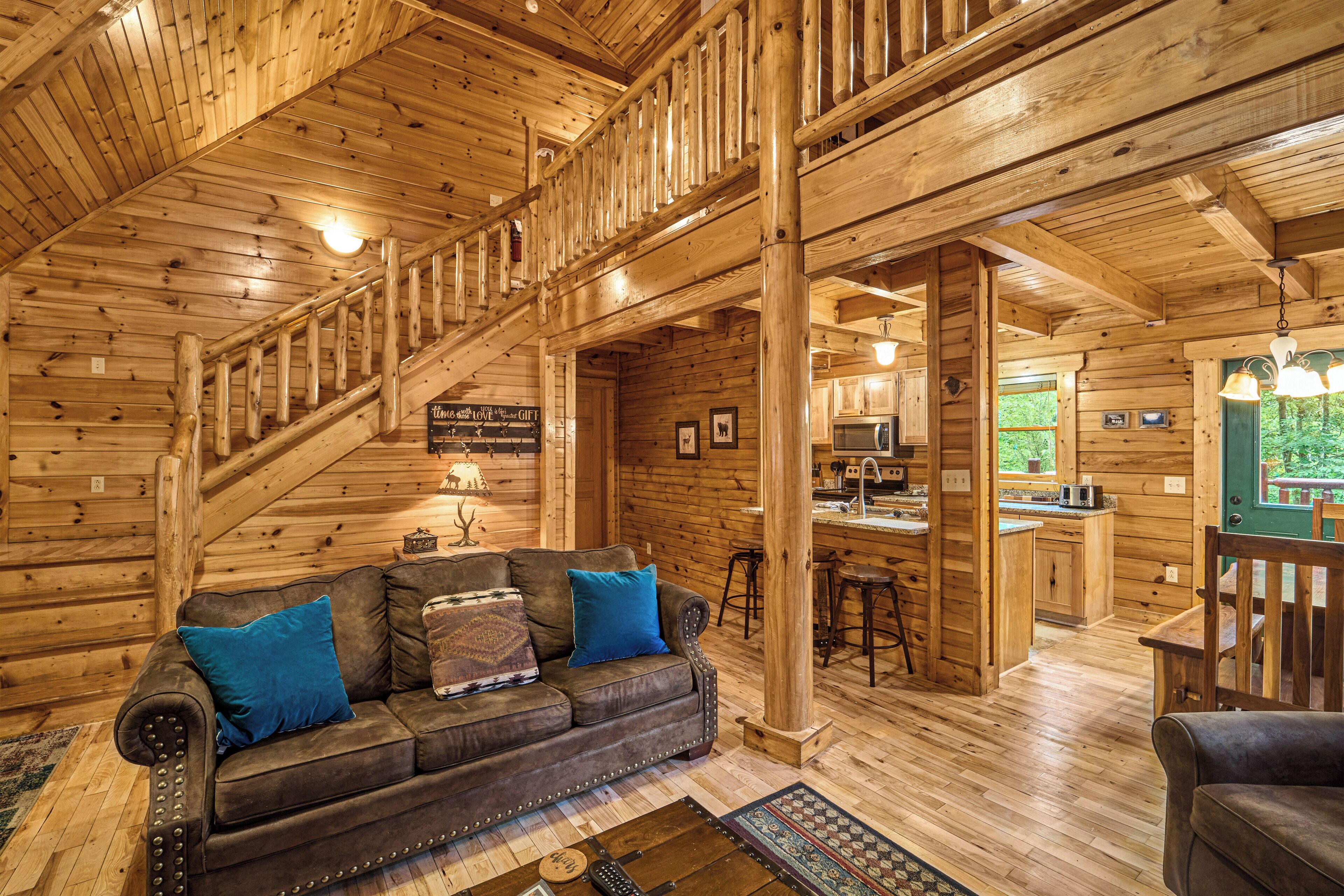 A wooden cabin interior with a staircase, a living room with a sofa and pillows, a kitchen area, and a dining table.