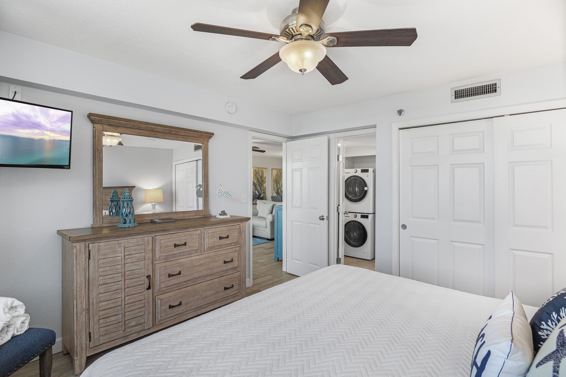 A bedroom with a bed, a dresser, a ceiling fan, a washing machine, and a door leading to another room.