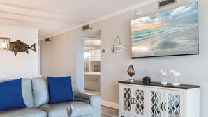 A living room with a grey sofa, a coffee table with decorative items, a white cabinet with glass doors, and a large painting of a beach scene on the wall. - 🌊Islander #509 – Where the Sand is the Softest & the Views Are Endless🏖️✨ (Destin)