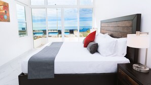 A bedroom with a large window overlooking a pool and beach, a bed with white bedding and gray and red pillows, and a bedside table with a lamp. - Oceanfront Villa Amor (Villas Corona)