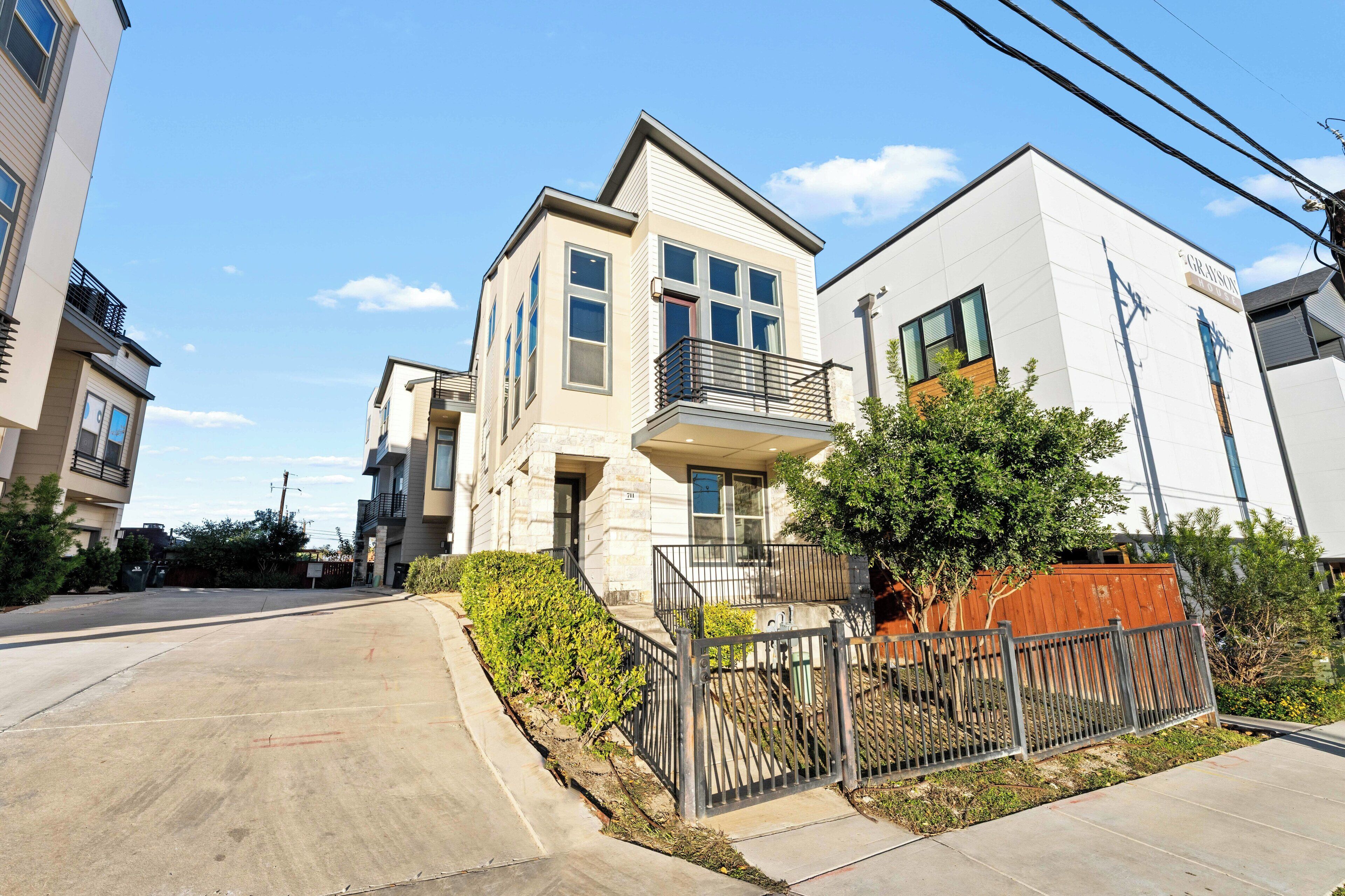 A modern two-story residential building with a balcony, a gated driveway, and a clear sky.