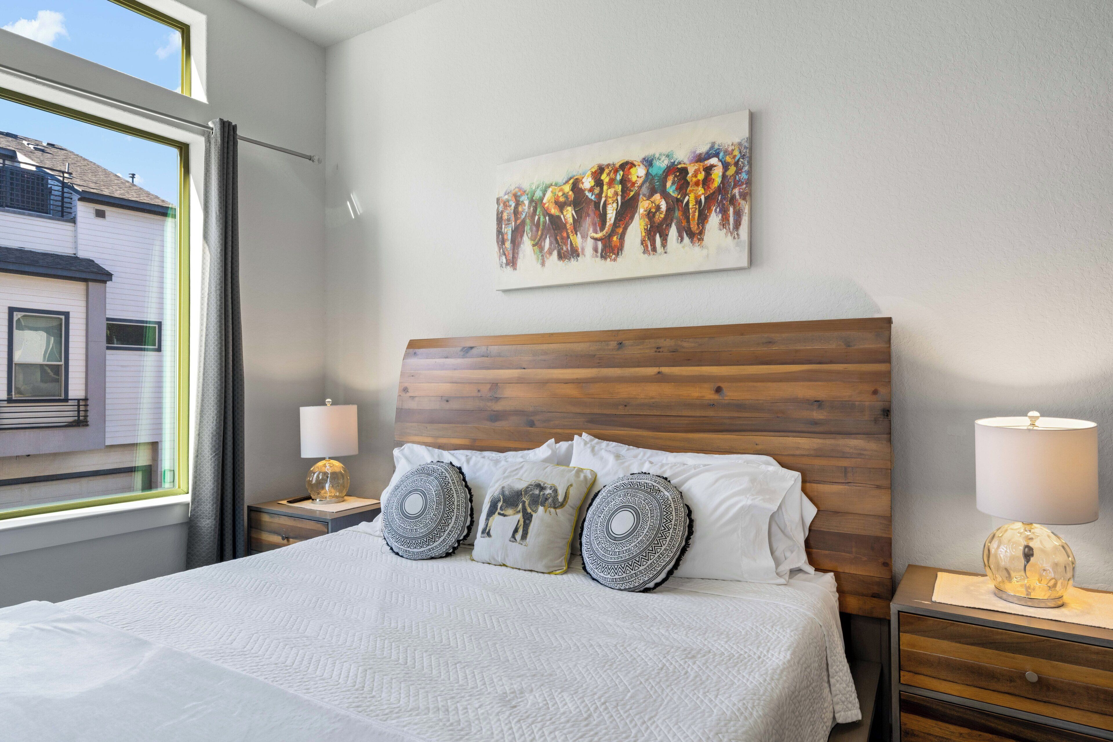 Una cama bien tendida con almohadillas estampadas, un cabecero de madera y un cuadro de un elefante colgado en la pared.
