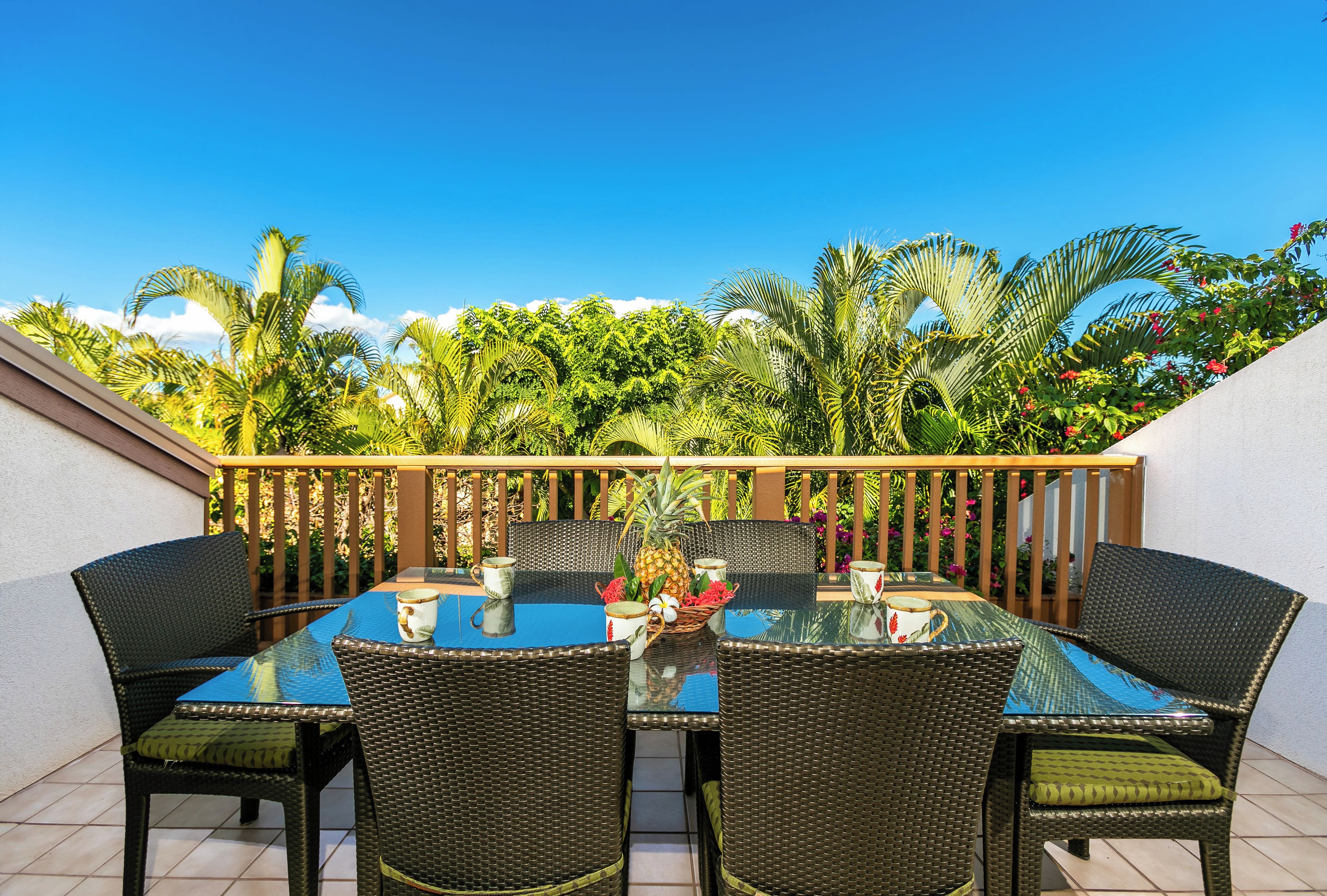 An outdoor dining area with wicker chairs and a glass table, surrounded by lush greenery and a wooden railing.