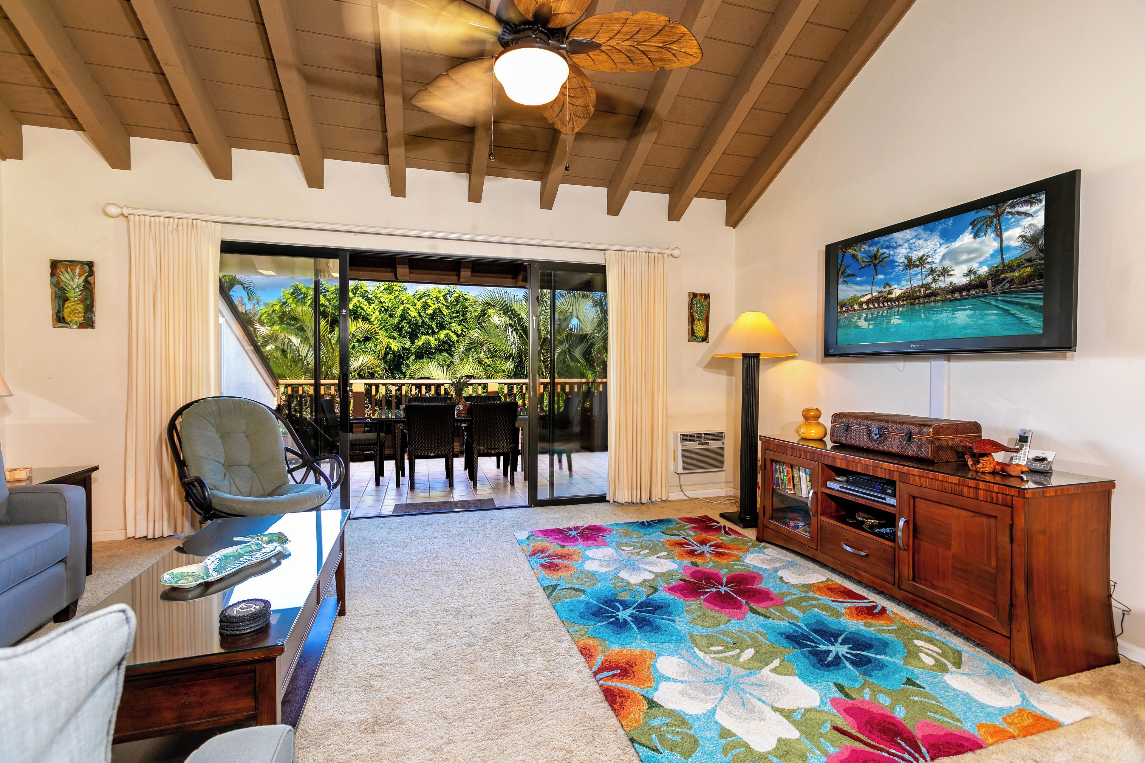 A room with a ceiling fan, a large window with curtains, a dining area with chairs and a table, a colorful rug, and a wooden cabinet with a television.