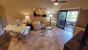 A living room with a dining table, a sofa, and a coffee table. There is a ceiling fan, a TV, and a window with blinds. - Partial Ocean View from private lanai, Queen Bed.....fully furnished condo (Kihei)