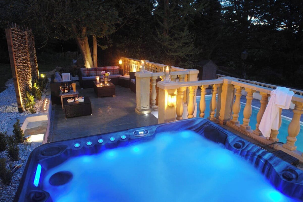 A hot tub with blue lighting, outdoor seating, and a poolside area with a wooden deck and railing.