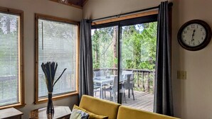 Ein Zimmer mit einem gelben Sofa, einem Tisch mit einer Vase und Blick auf eine Terrass mit Stühlen und einer Uhr an der Wand.