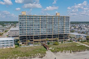 A multi-story hotel with a parking garage, located near a beach with a sandy path leading to the entrance.