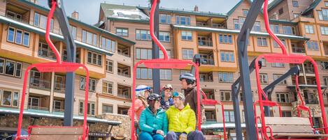Un telesilla con pasajeros, un edificio de varios pisos con balcones y suelo cubierto de nieve.