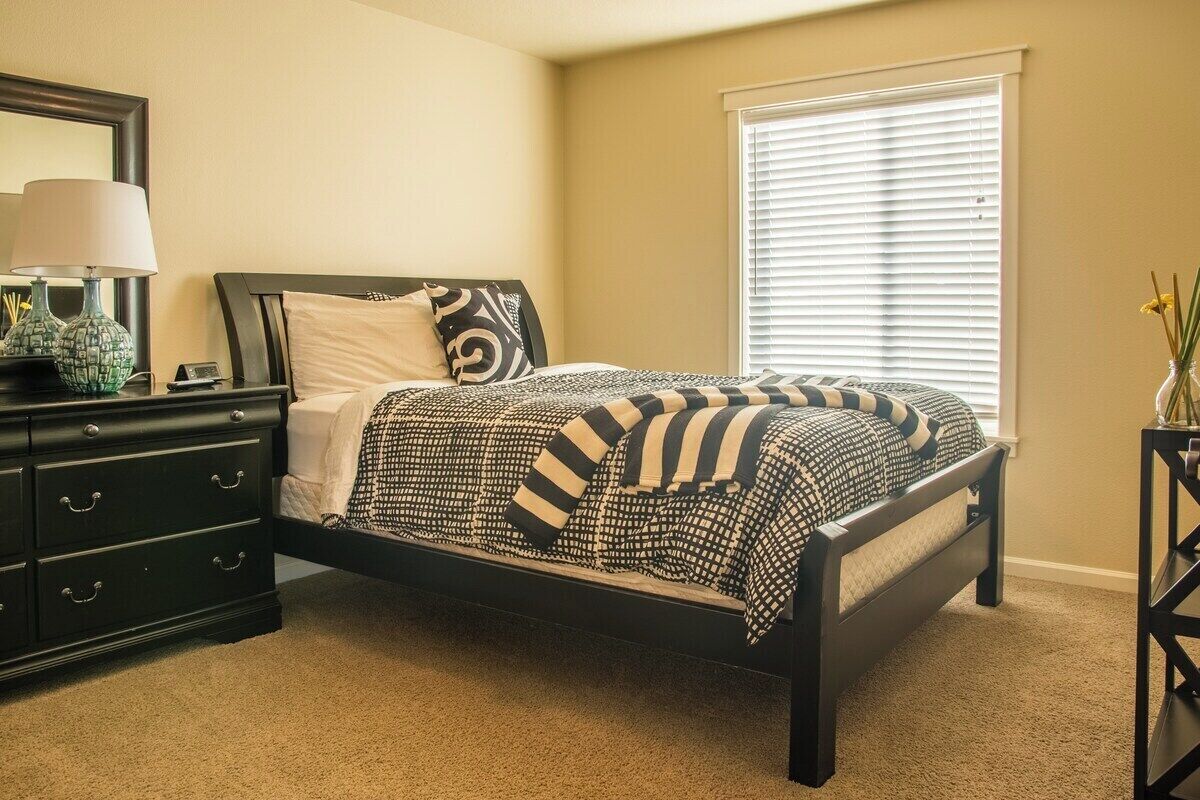 A bedroom with a bed, a nightstand, a dresser, a window with blinds, and a vase with flowers.