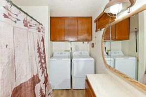 A compact laundry room with two washing machines, a wooden cabinet above, and a mirror with a wooden frame.