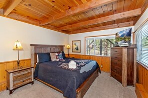 Une chambre avec un plafond en bois, un lit recouvert d’une couvre-lit bleu foncé, une table de chevet avec une lampe, une commode et un téléviseur posé sur un meuble.