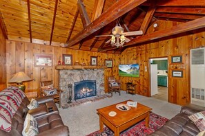 A cozy log cabin interior with a fireplace, leather sofas, a wooden ceiling, and framed pictures on the walls.