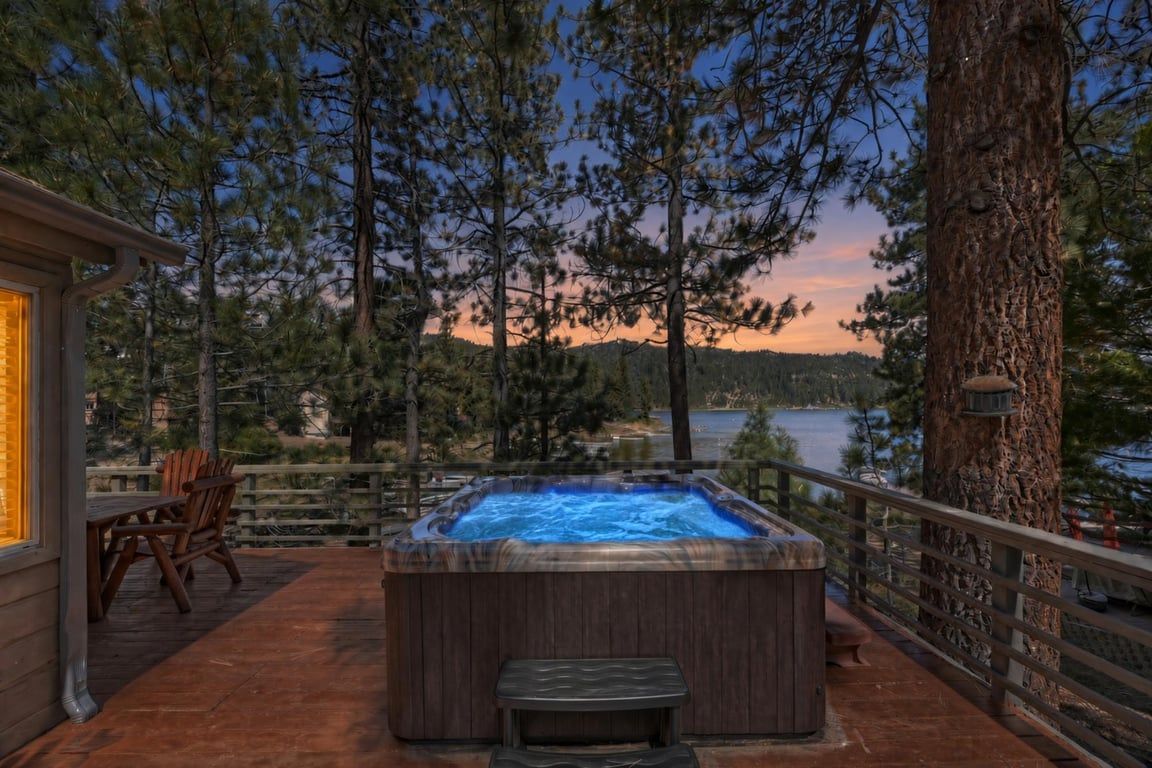 Een hottub op een houten terras, met stoelen, met uitzicht op een meer, omringd door bomen.