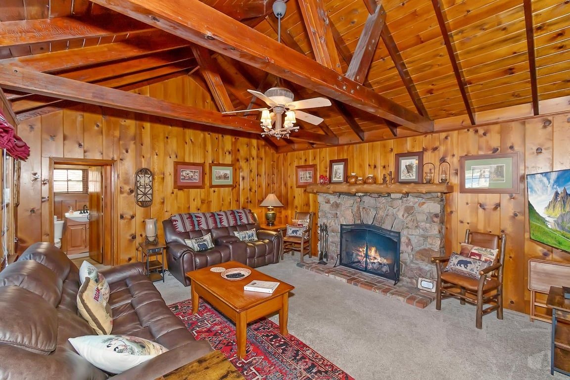 A log cabin interior with a fireplace, leather sofa, wooden coffee table, and a ceiling fan.