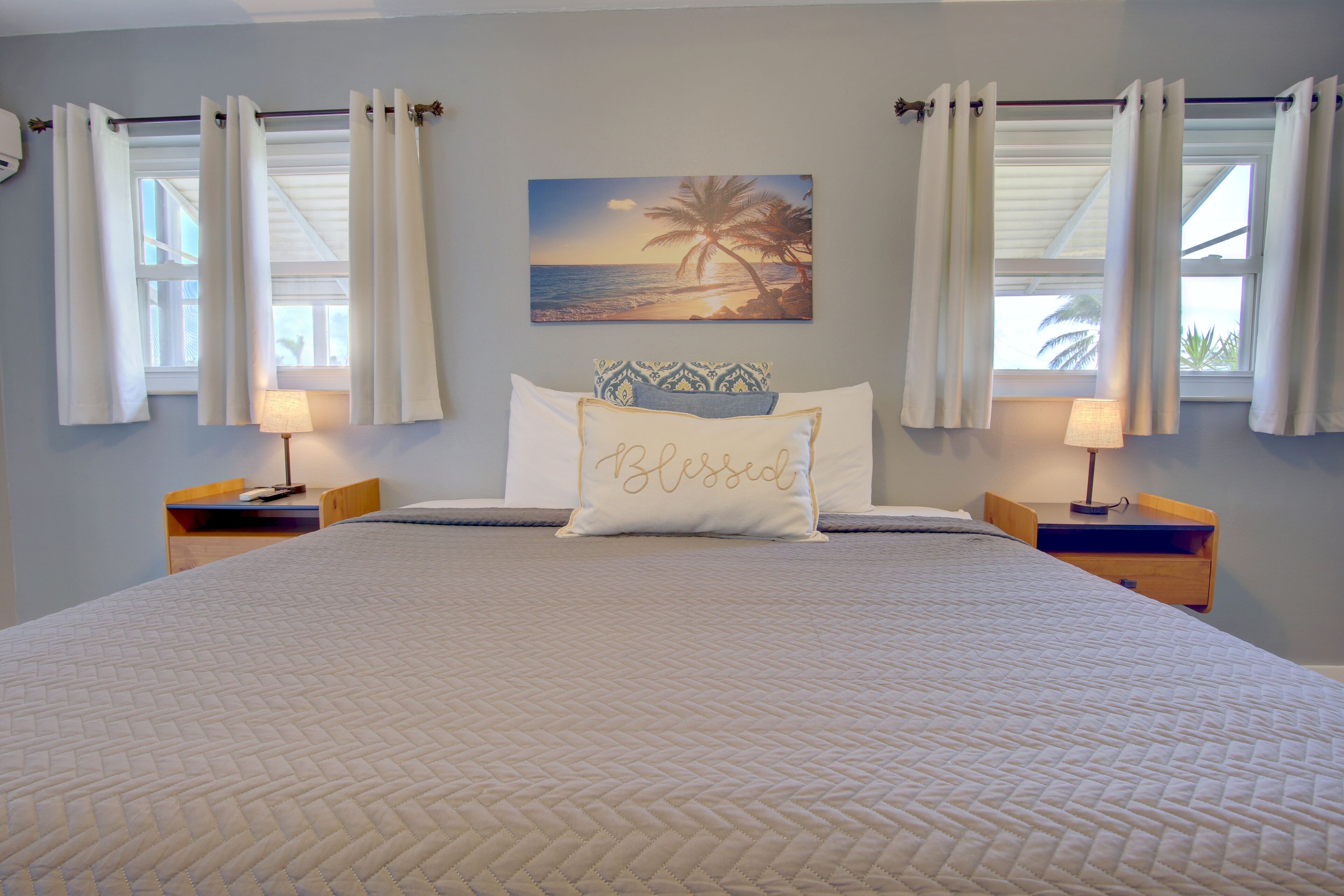 A bedroom with a large bed, two bedside tables with lamps, a wall-mounted painting of a beach scene, and a pillow with the word "Blessed" on it.
