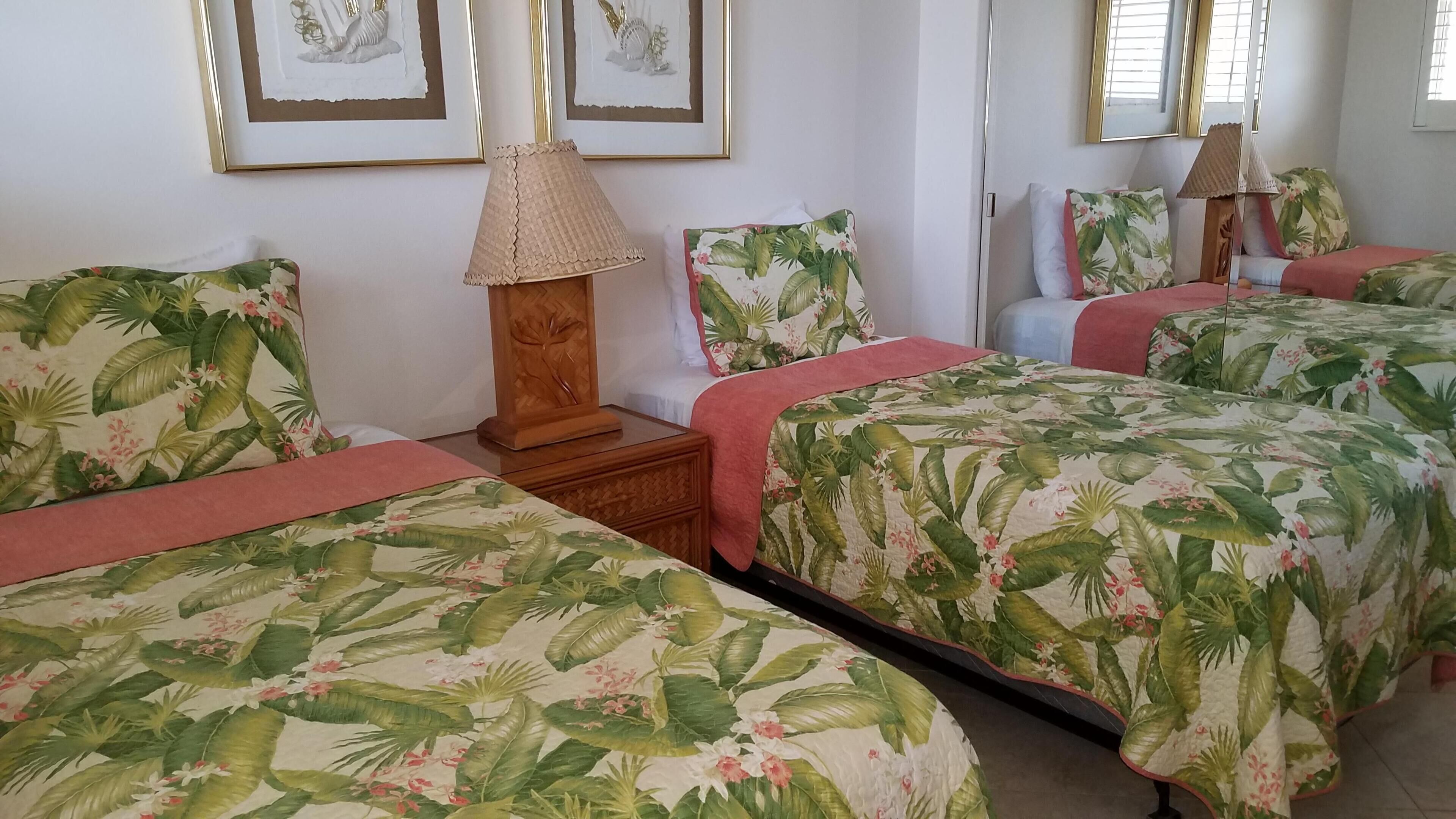 Une chambre d’hôtel avec trois lits, chacun recouvert d’une couvre-lit à motifs floraux, une table de chevet en bois avec une lampe, et un mur orné de tableaux encadrés.