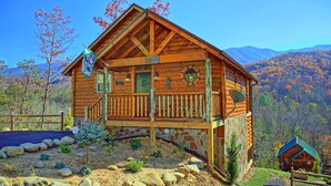 Una cabaña de troncos con base de piedra, una terraza de madera y montañas al fondo.