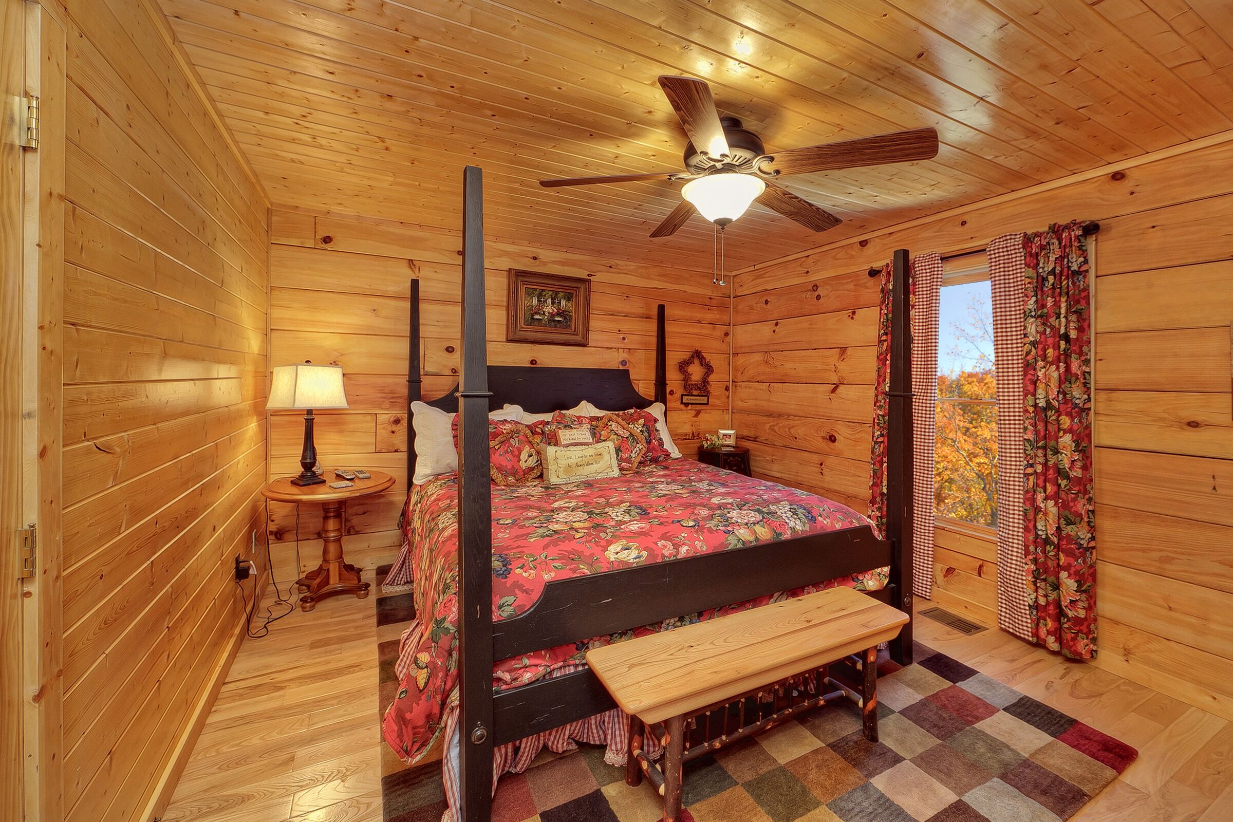 A wooden cabin bedroom with a four-poster bed, a ceiling fan, a nightstand, and a window with curtains.