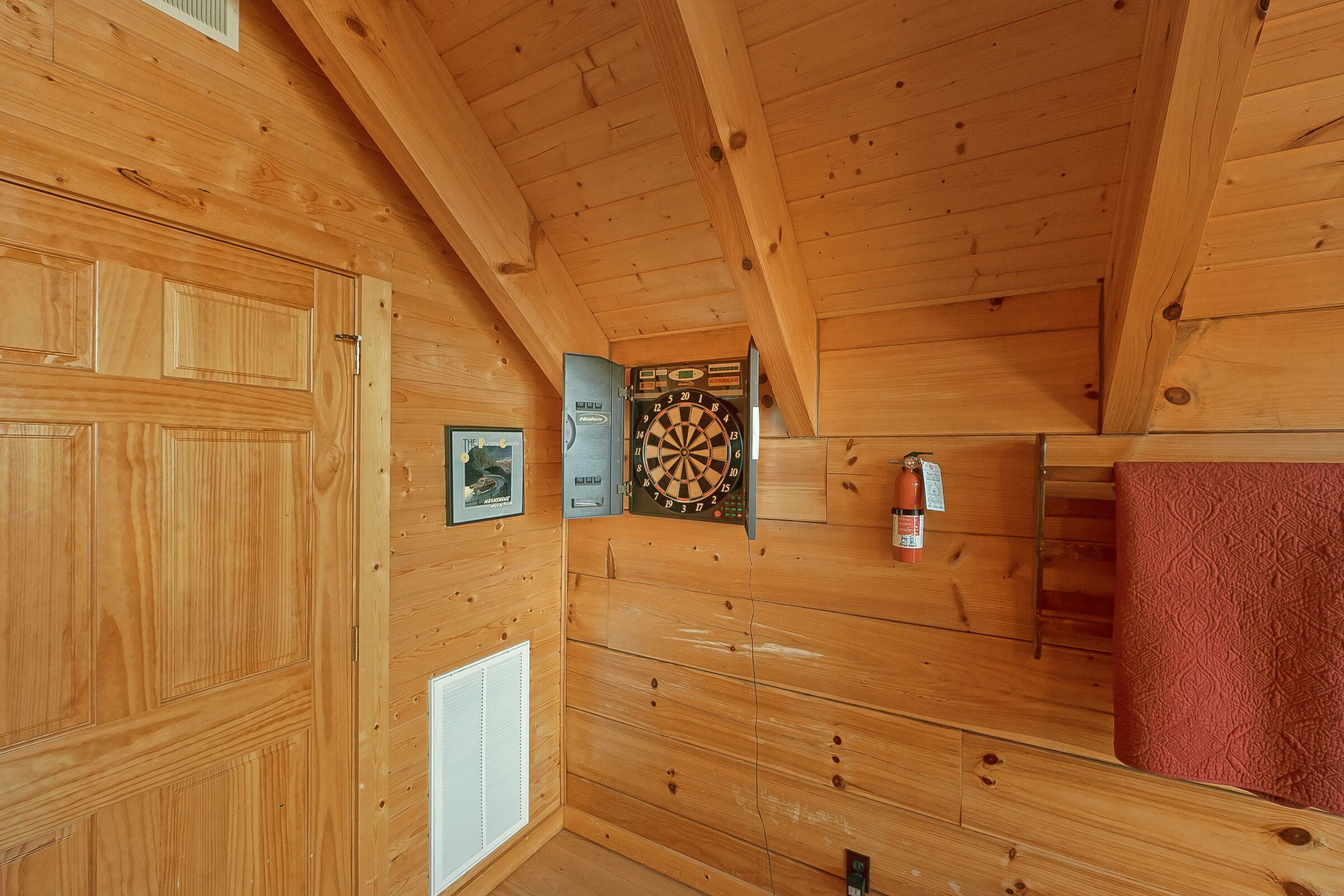 A wooden interior with a dartboard, framed pictures, a fire extinguisher, and a red textured wall.