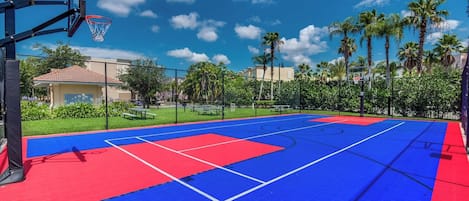 Una cancha de baloncesto al aire libre con superficie azul y roja, rodeada por una valla, palmeras y un cielo despejado.