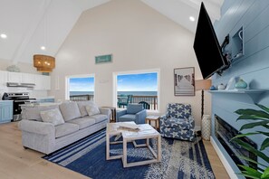 A living room with a sofa, armchair, and coffee table, a television mounted on the wall, and a view of the ocean through large windows.