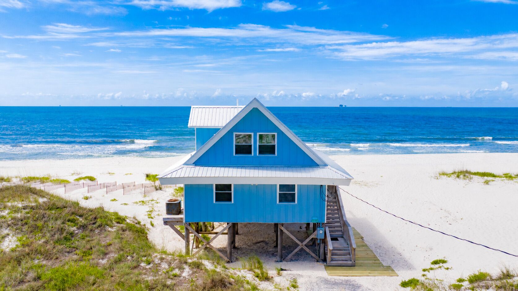 Una casa de playa azul con una escalera que lleva a una terraza, ubicada en una playa arenosa con el océano al fondo.