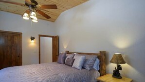 A bedroom with a wooden ceiling fan, a bed with patterned bedding, a nightstand with a lamp, and a wooden door.