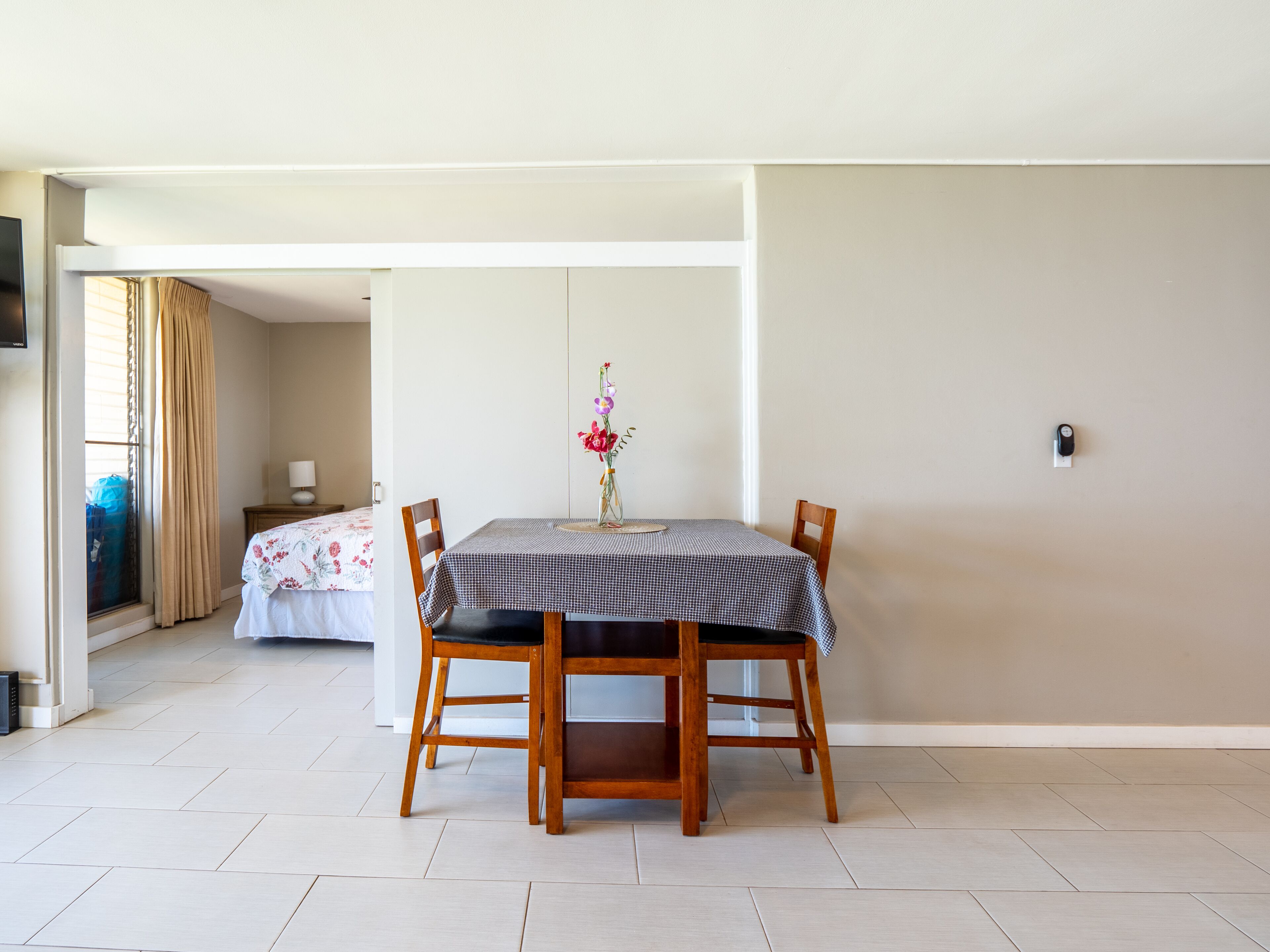 A dining area with a table set for two, a vase with flowers, and a bed in the background.